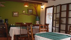 Un espace repas avec des chaises et des tables en bois, un mur vert orné d’une fresque représentant un arbre, et une fenêtre avec des barreaux.