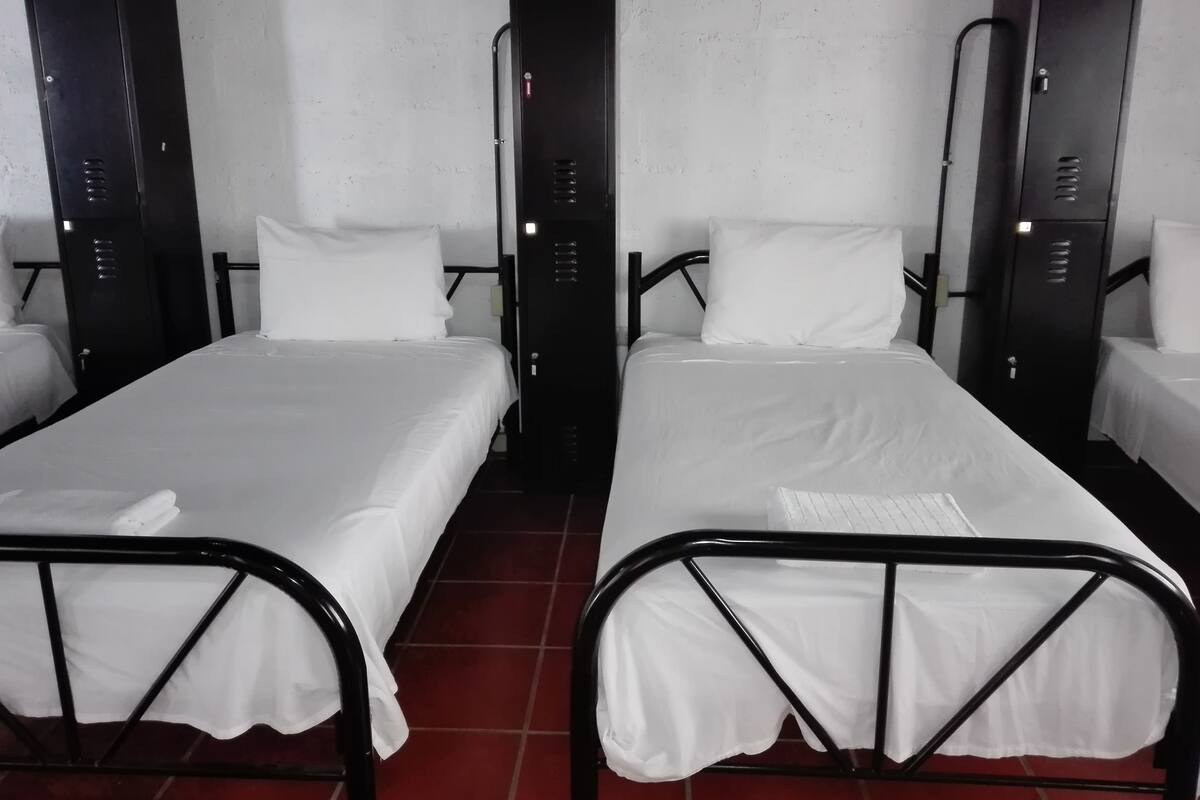 Une chambre avec deux lits simples, chacun doté d’une structure en métal noir et de draps blancs. De chaque côté des lits, on trouve des casiers noirs.