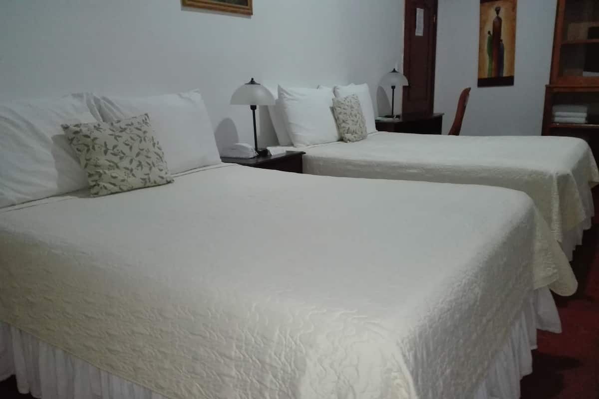 Une chambre d’hôtel avec deux lits, chacun agrémenté de draps blancs et d’un oreiller à motifs floraux. Une table de chevet avec une lampe se trouve entre les deux lits.