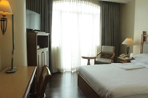 A hotel room with a bed, a desk, a chair, a small table, a lamp, a TV, and a window with curtains. - The Grand Dame Hotel (Iloilo)