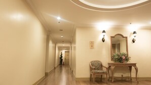 A hotel hallway with a wooden floor, a chair, a console table with a mirror and a flower arrangement, and a number sign on the wall. - The Grand Dame Hotel (Iloilo)