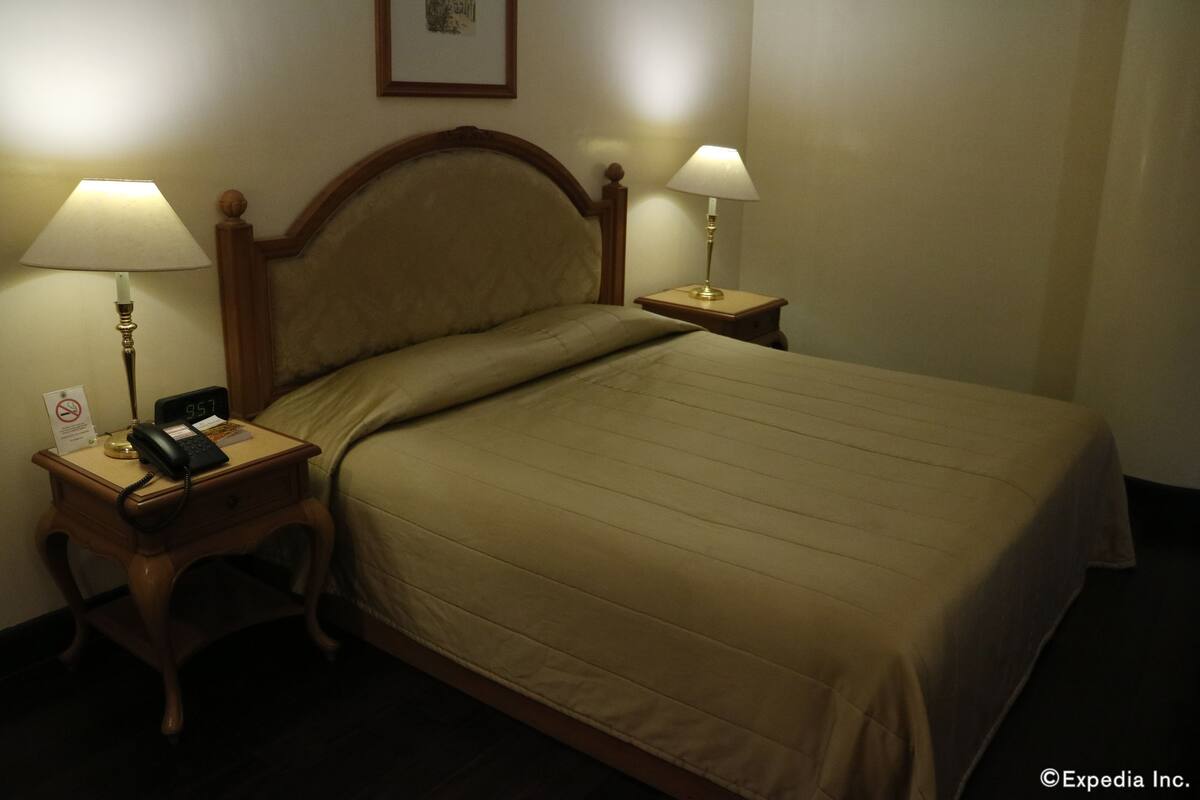 A hotel room with a bed, two bedside lamps, a nightstand with a phone, and a framed picture on the wall.