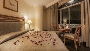 A hotel room with a bed, a bedside table with a lamp, a chair, and a view of the city through a large window. - The Grand Dame Hotel (Iloilo)
