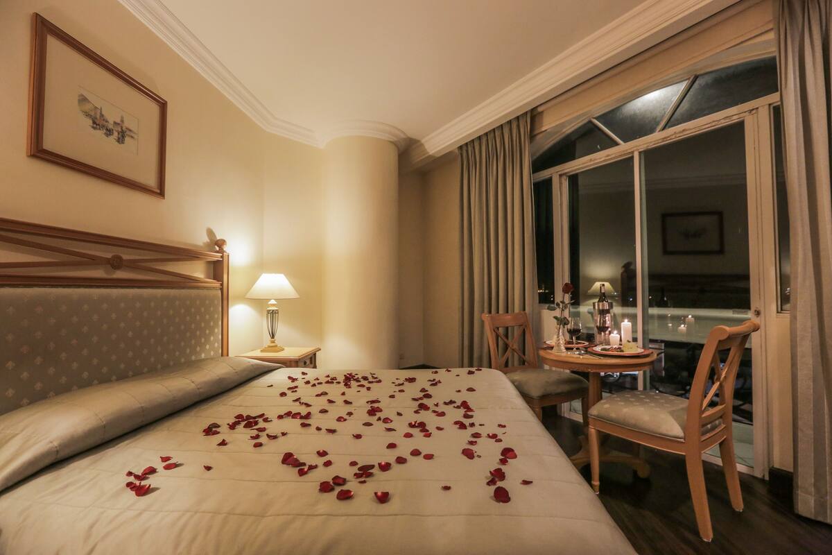 A hotel room with a bed, a bedside table with a lamp, a chair, and a view of the city through a large window.