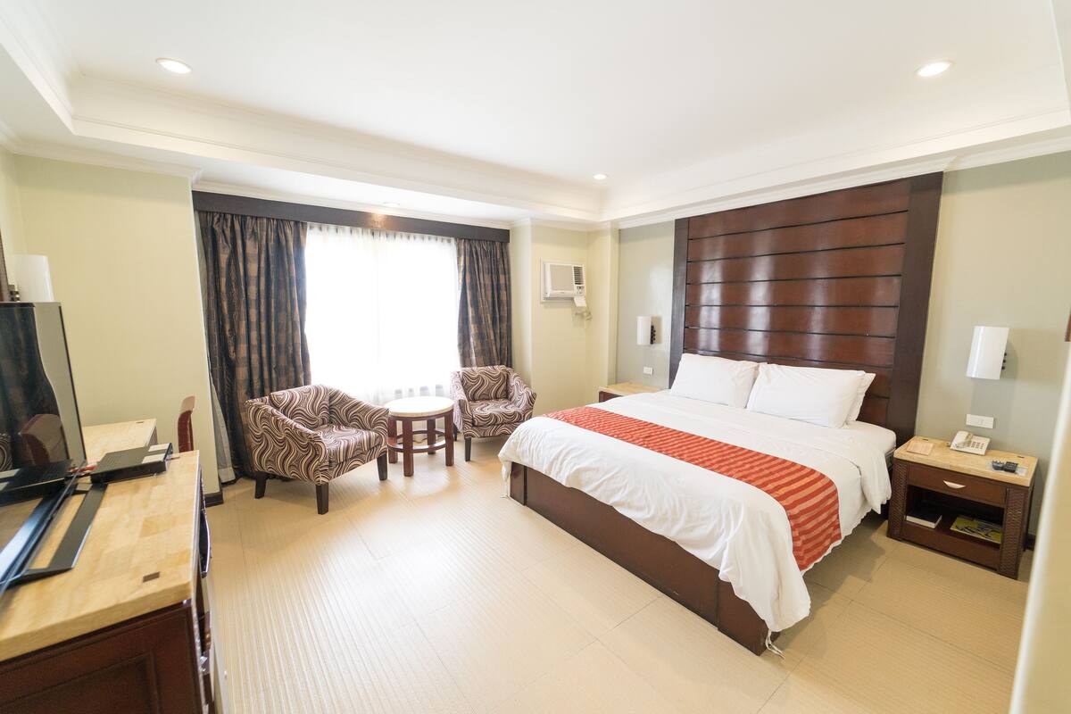 Een hotelkamer met een bed, een bureau met een computer, twee fauteuils, een kleine tafel en een raam met gordijnen.