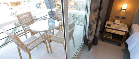 A hotel room with a balcony, wicker furniture, a glass table, a bedside table with a lamp, and a bed.