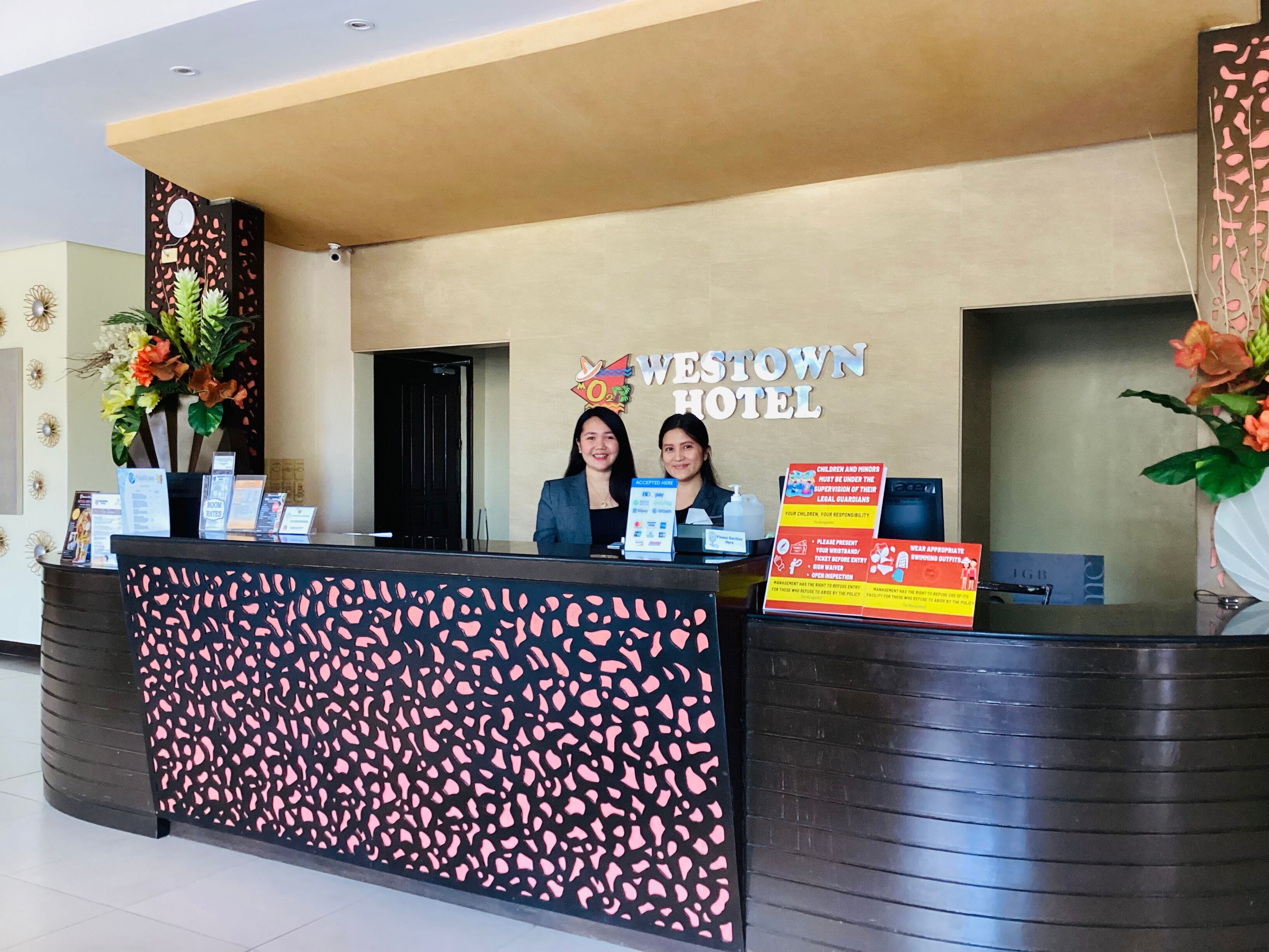 A reception desk at Westown Hotel with a modern design, including a patterned backdrop and a signboard with hotel name and amenities.