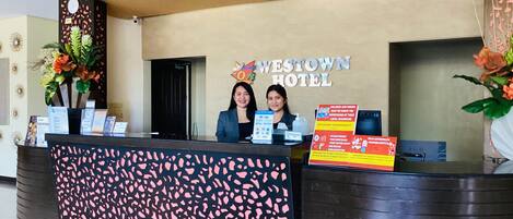 A reception desk at Westown Hotel with a modern design, including a patterned backdrop and a signboard with hotel name and amenities.