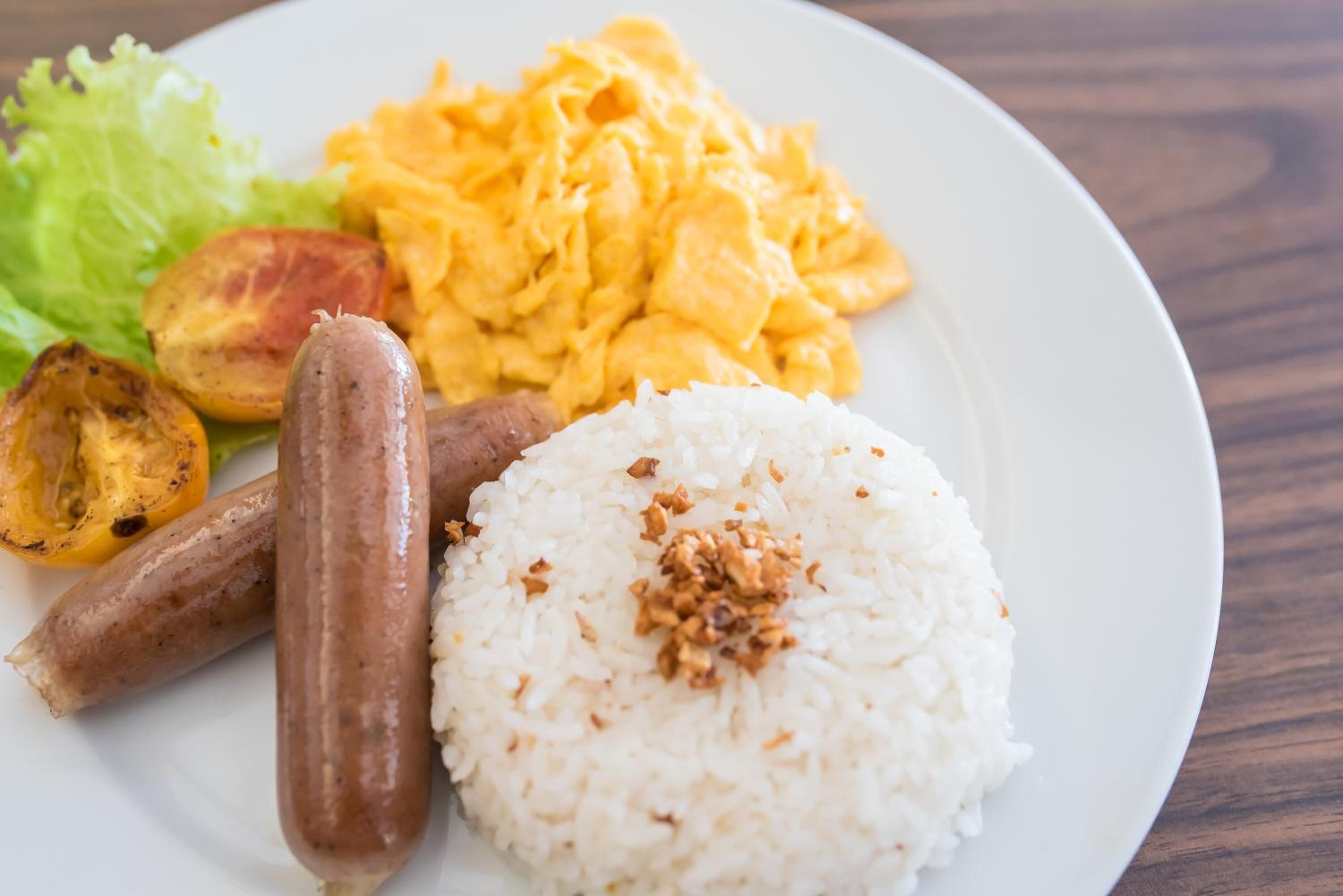 A plate with white rice topped with fried onions, two grilled sausages, scrambled eggs, and a side of lettuce and roasted tomatoes.