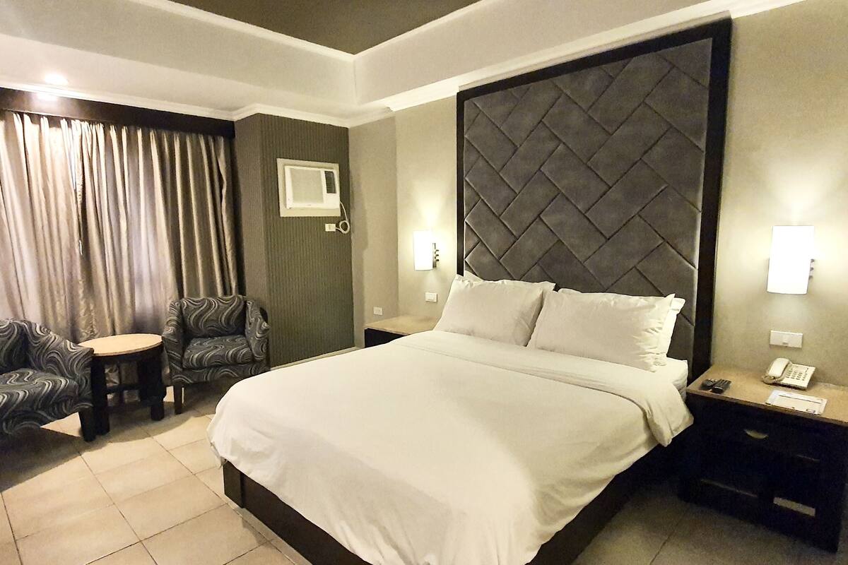 Een hotelkamer met een groot bed, twee fauteuils, een tafeltje, een nachtkastje met telefoon en een raam met gordijnen.