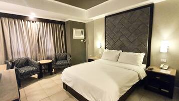 Habitación de hotel con una cama grande, dos sillones, una mesita, un mesillón con teléfono y una ventana con cortinas.