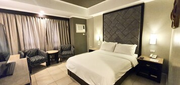 A hotel room with a large bed, two armchairs, a small table, a nightstand with a phone, and a window with curtains.