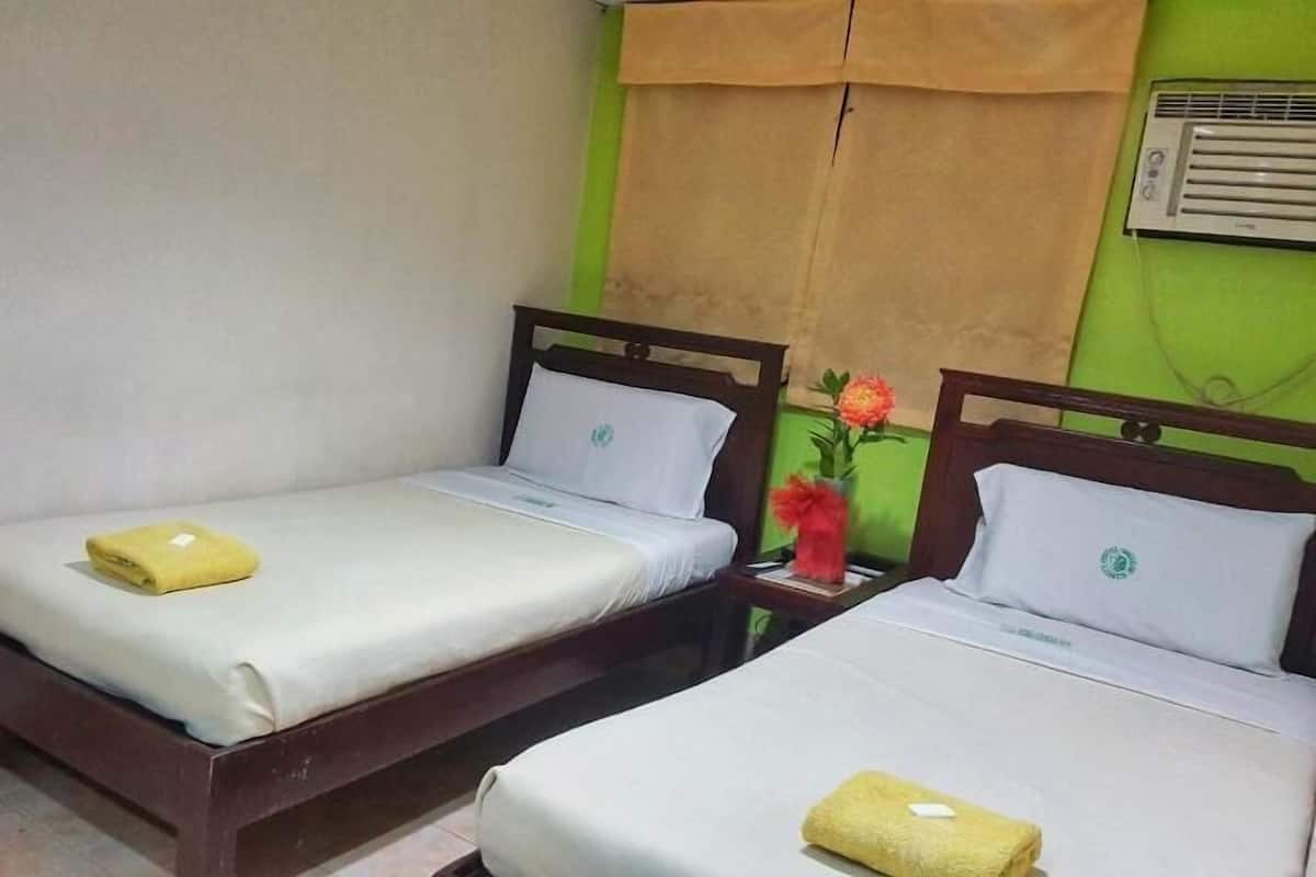 A hotel room with two single beds, each with a wooden headboard, white bedding, and a yellow towel. A green wall and a window with a brown curtain are visible.