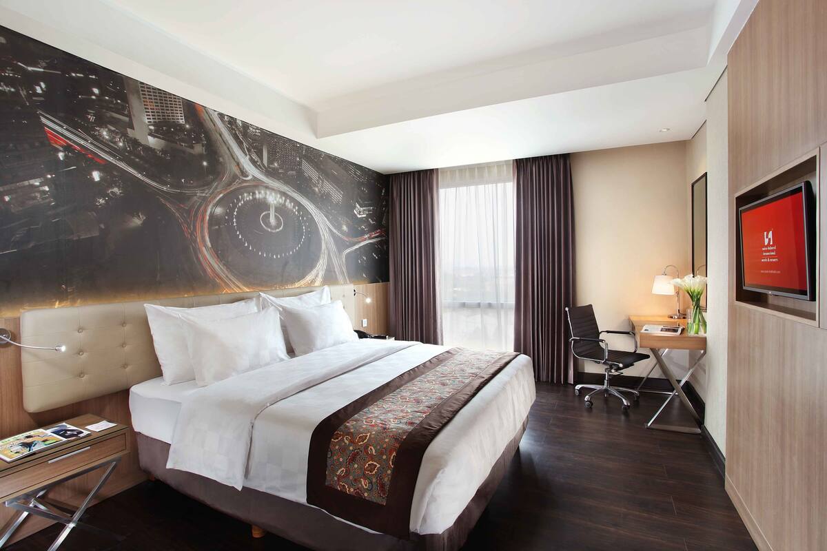 Une chambre d’hôtel avec un grand lit, un bureau avec une chaise, une télévision et une fresque représentant un paysage urbain sur le mur.