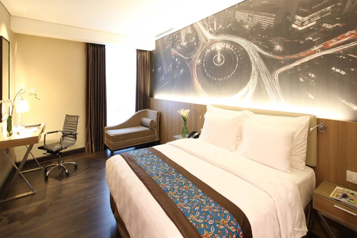 Une chambre d’hôtel avec un grand lit, un bureau avec une chaise, une petite table avec un vase et un mur orné d’une fresque représentant un paysage urbain.