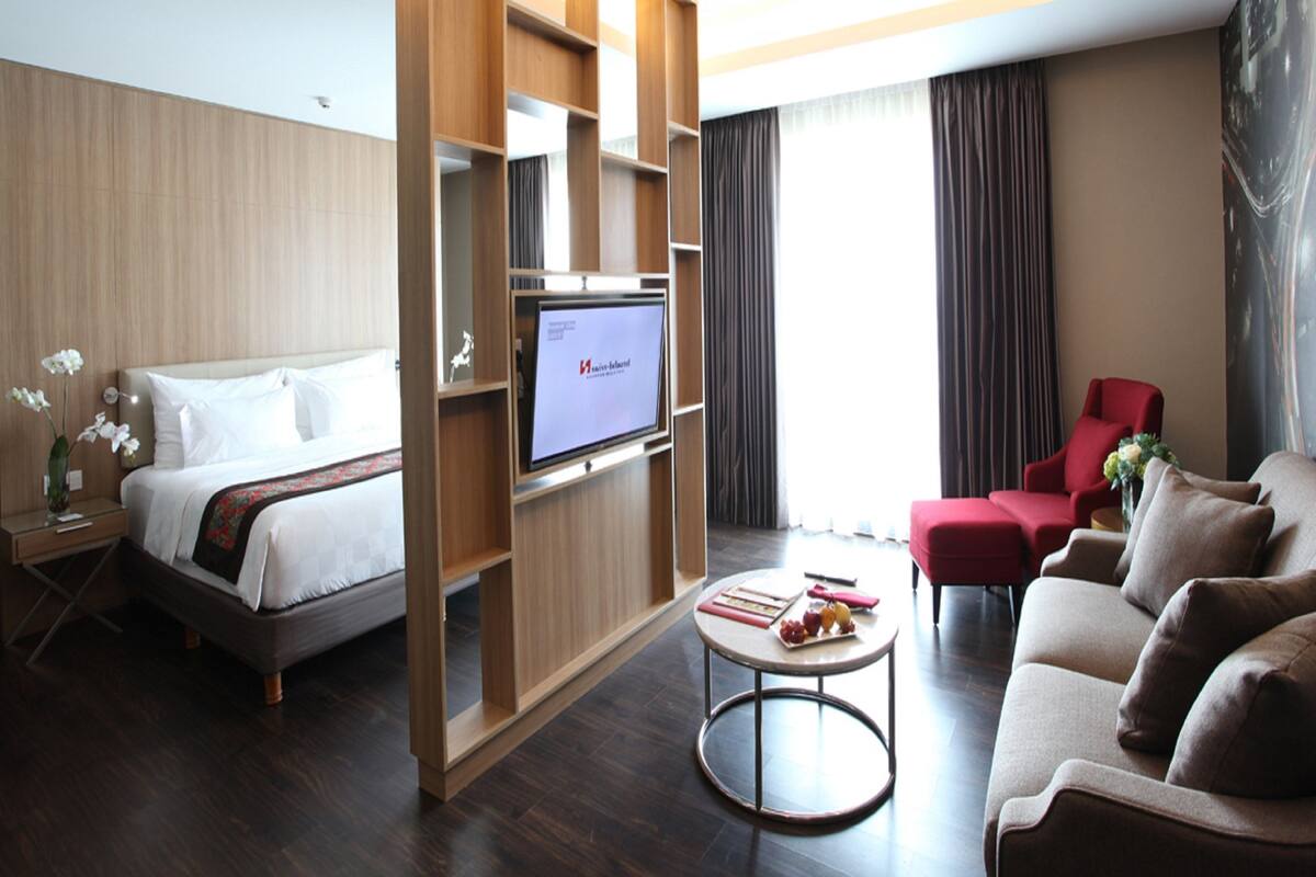 Une chambre d’hôtel avec un lit, une télévision, un fauteuil rouge, un canapé et une petite table sur laquelle se trouvent des fruits.