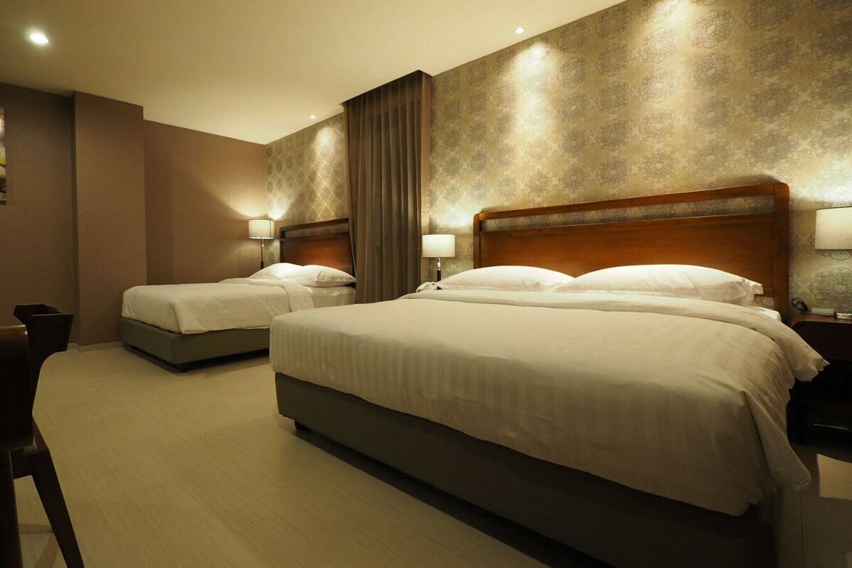 Habitación de hotel con dos camas individuales y una cama más grande, todas con ropa blanca. Hay mesitas de noche con lámparas, una silla y un televisor montado en la pared.