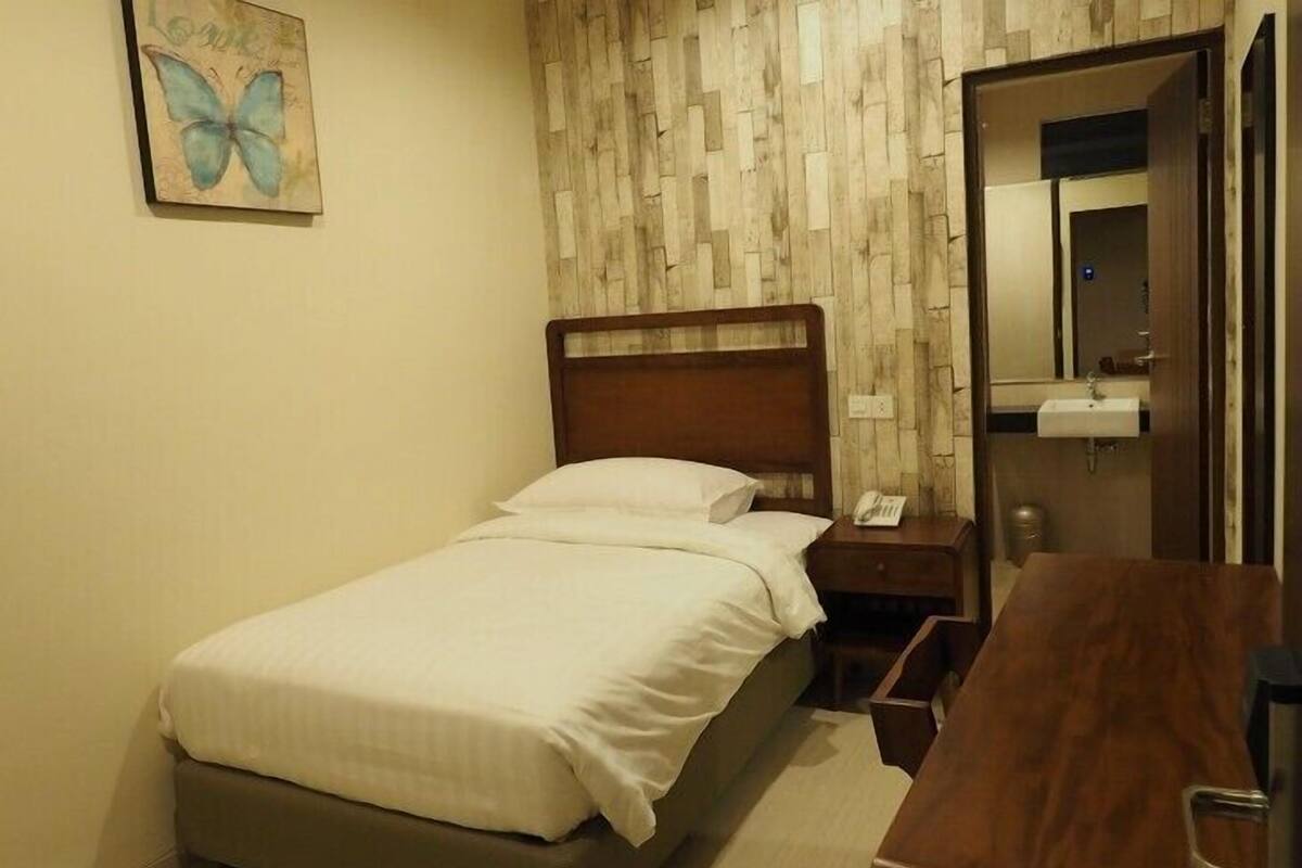 Un dormitorio con cabecera de madera, cama con ropa de cama blanca, mesita de noche con un teléfono, cómoda de madera, espejo y una obra de arte en la pared de una mariposa.