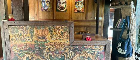 Un mostrador de recepción rústico de madera con intrincados diseños florales, un estante de madera con libros y un abanico, y un objeto decorativo sobre el estante.