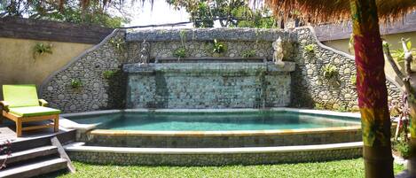 Una piscina con un muro de piedra y escalones que llevan a ella, rodeada de césped y una estructura cubierta de paja.