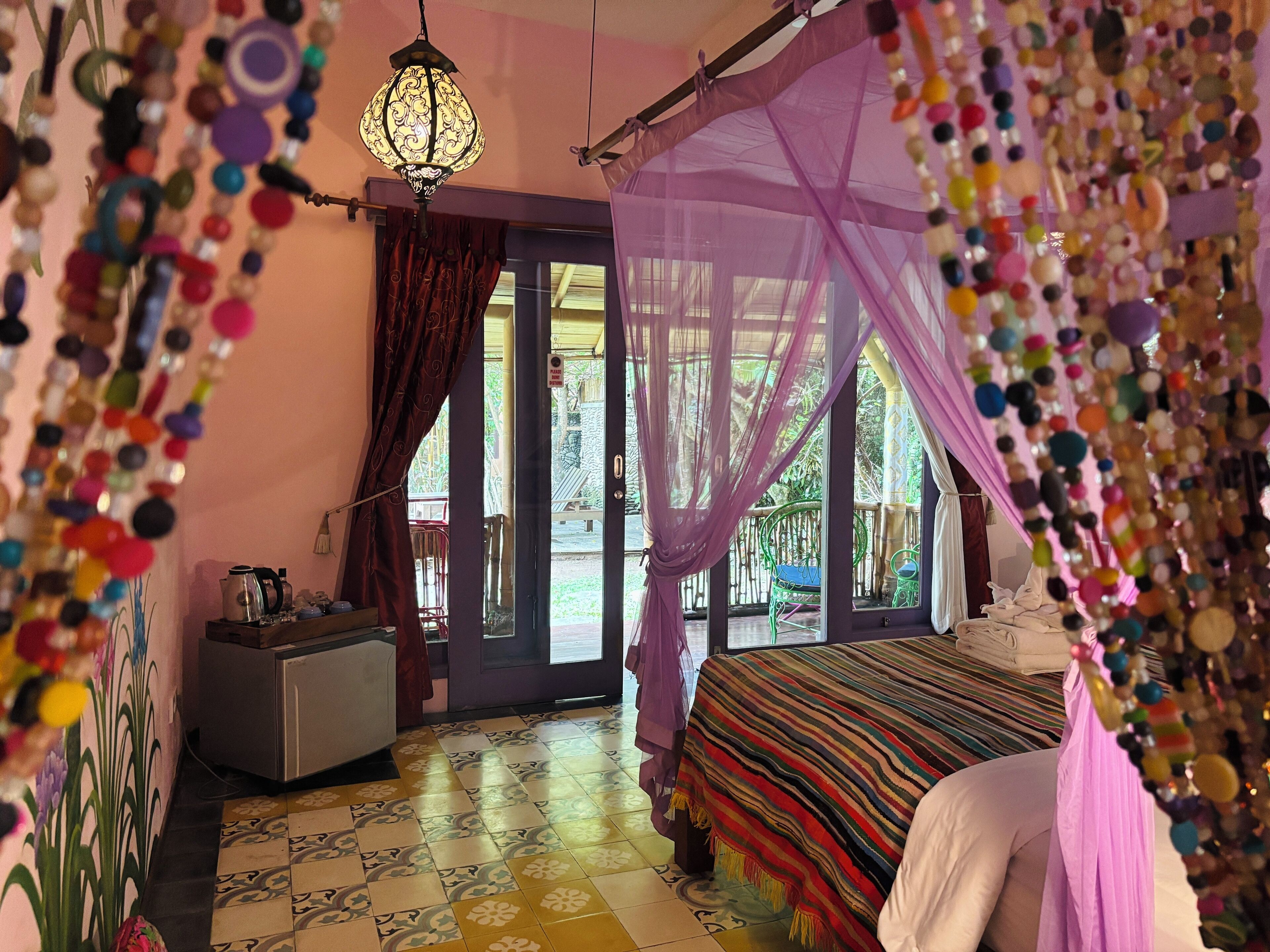 A room with a bed, a desk, and a chair. There is a window with curtains and a view of the outside.