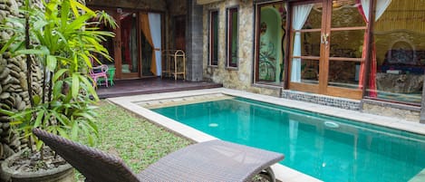 Una piscina con una tumbona, un muro de piedra y una estructura con techo de paja.