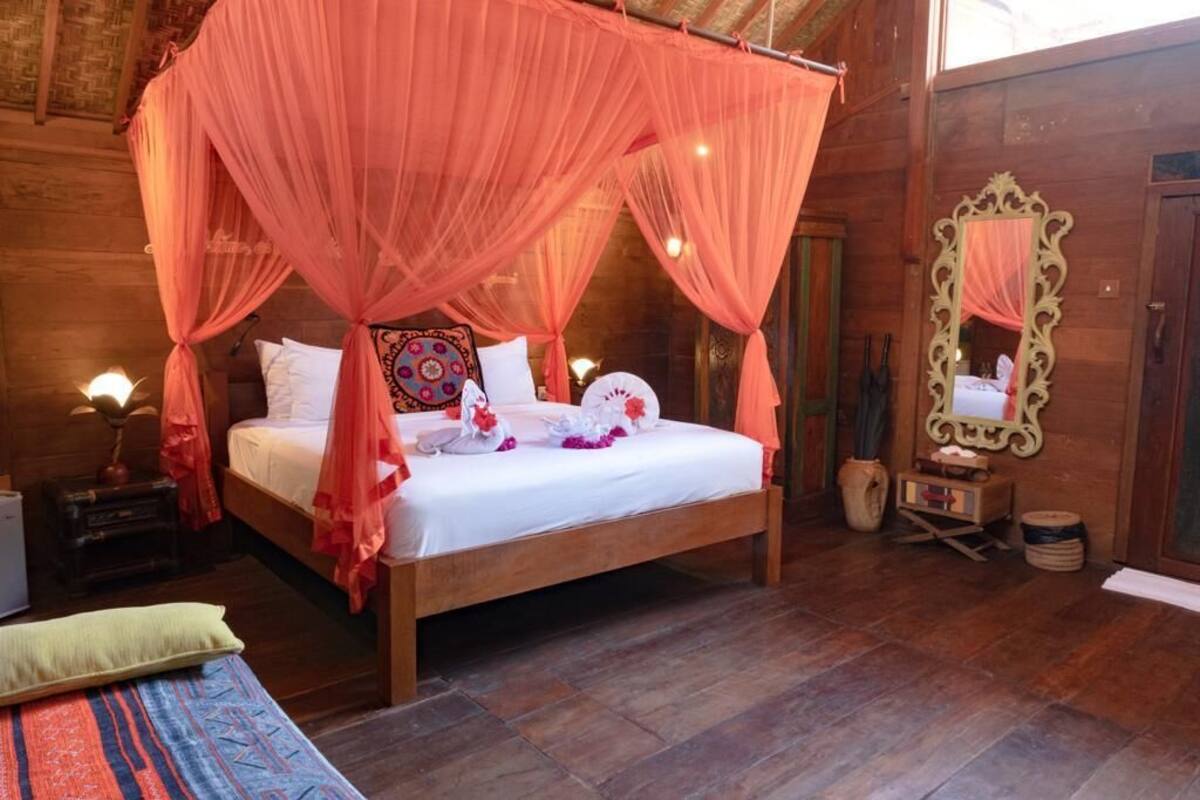 A wooden room with a bed under a canopy, a mirror, and a nightstand with a lamp.