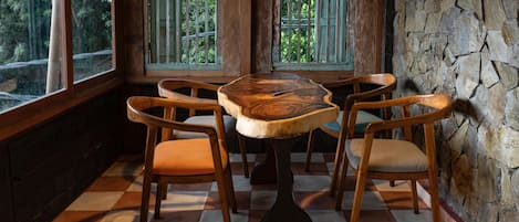 A dining area with a wooden table and chairs, large windows with bars, and a stone wall.