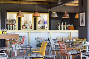 Zona de bar con sillas y mesas coloridas, estanterías con botellas y lámparas colgantes.