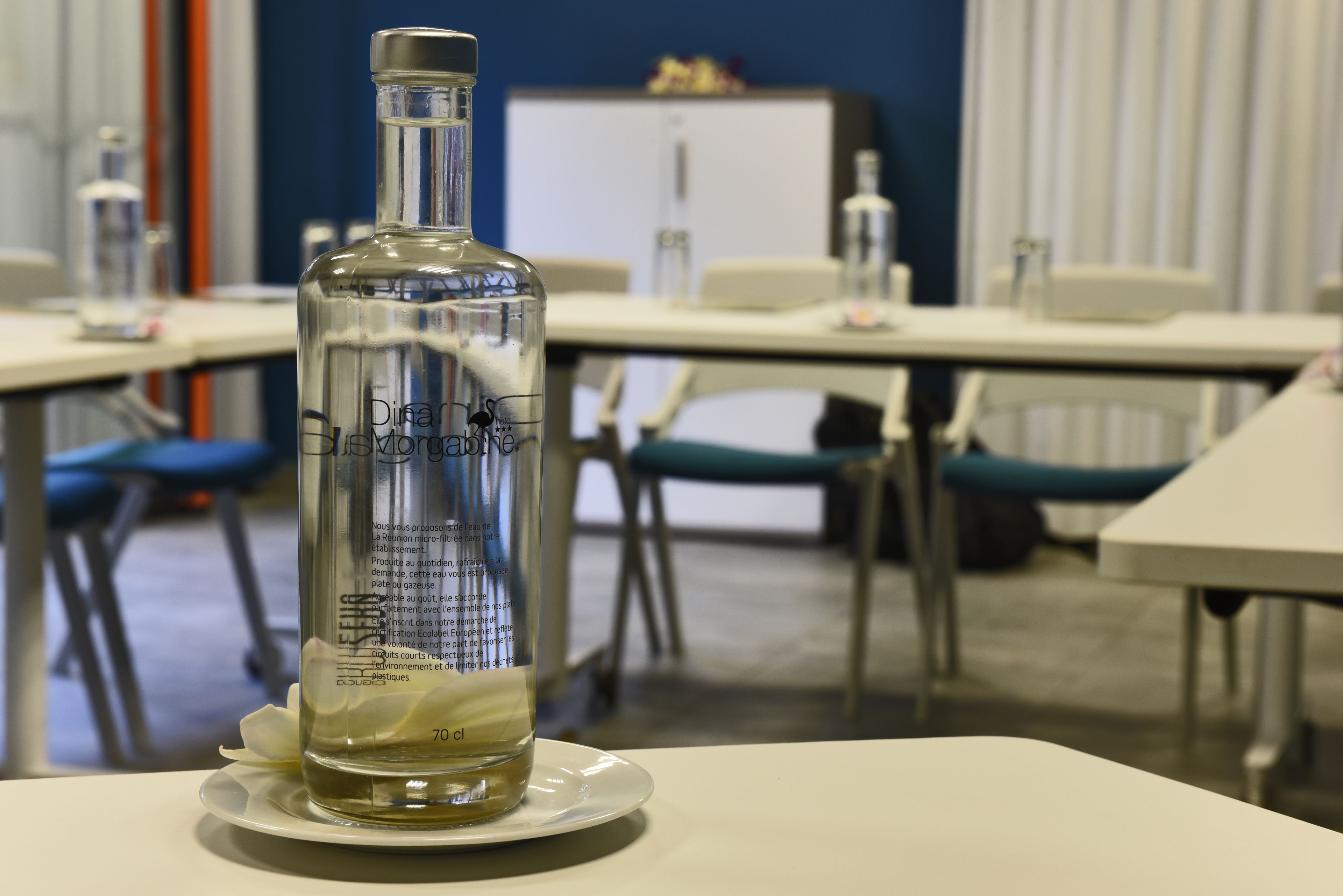 A bottle of 'Dina de Vries' on a white plate in a room with a blue wall, white furniture, and a whiteboard in the background.