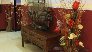 A lobby with a wooden table displaying a ship model, a vase with flowers, and a wall-mounted lamp. - Hollywood Hotel (Jakarta)