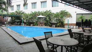 An outdoor swimming pool with a covered seating area, surrounded by palm trees and air conditioning units.