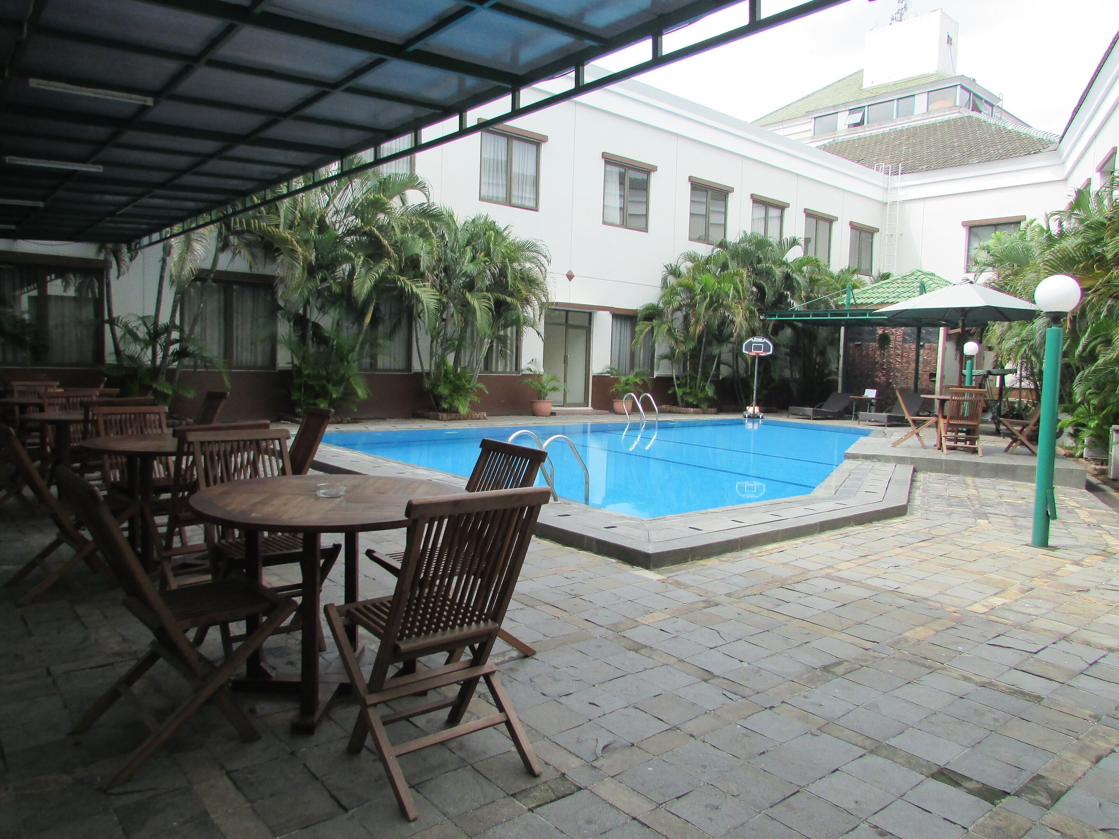 An outdoor pool area with wooden tables and chairs, surrounded by potted plants and a building in the background.