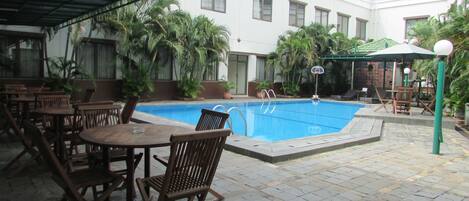 An outdoor pool area with wooden tables and chairs, surrounded by potted plants and a building in the background.