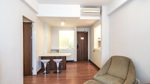 A hallway with a wooden floor, a seating area with a patterned armchair, and a kitchenette with a sink and two stools.