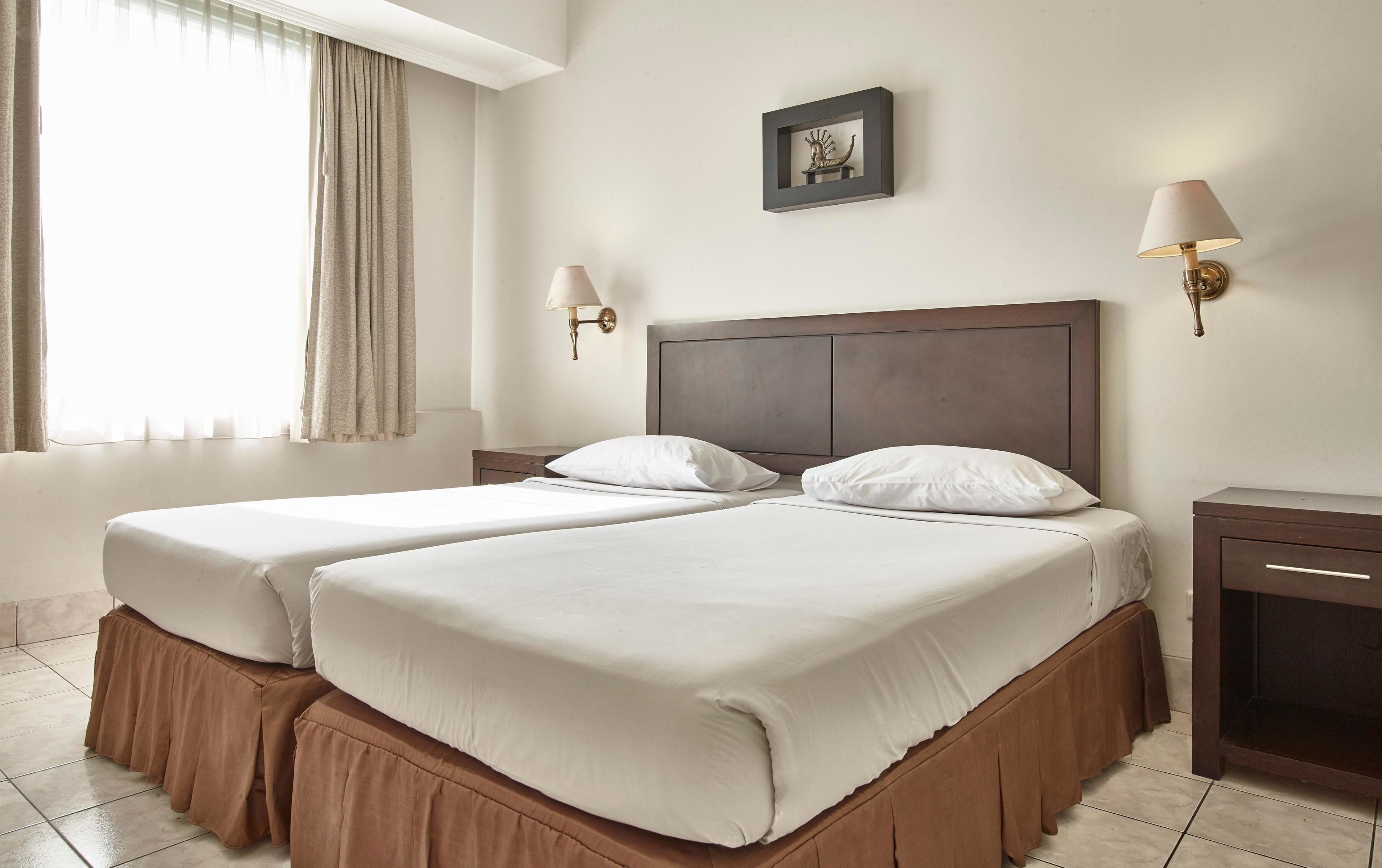 A hotel room with two beds, a nightstand, a lamp, and a window with curtains.