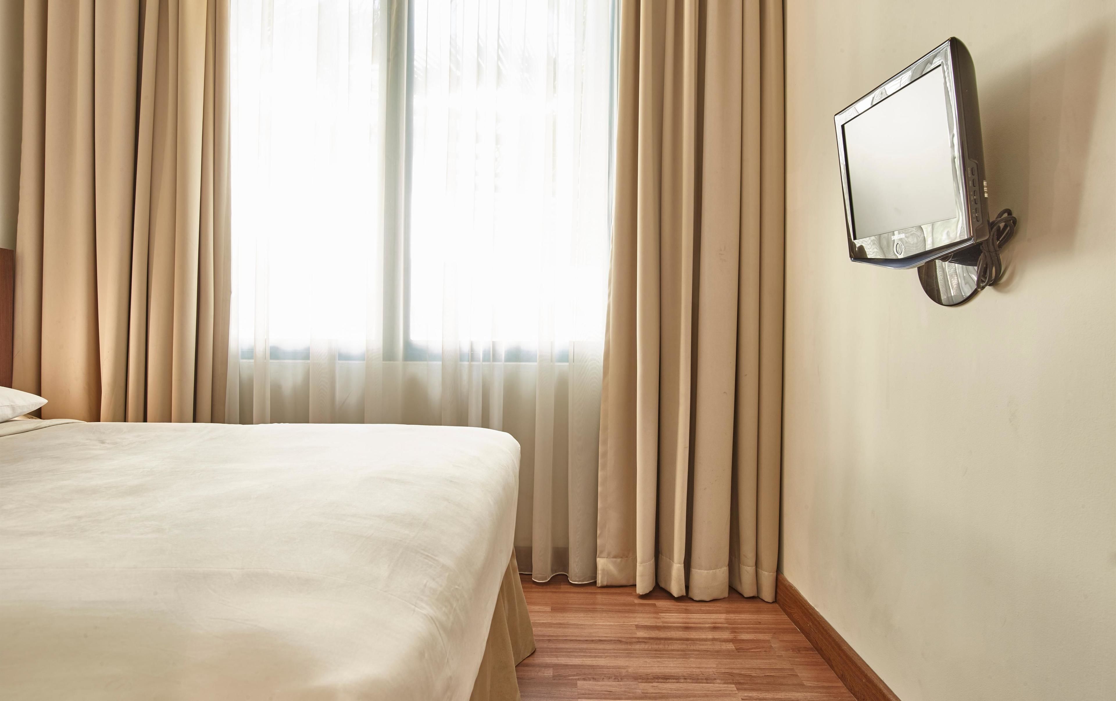 A hotel room with a bed, curtains, a wall-mounted TV, and wooden flooring.