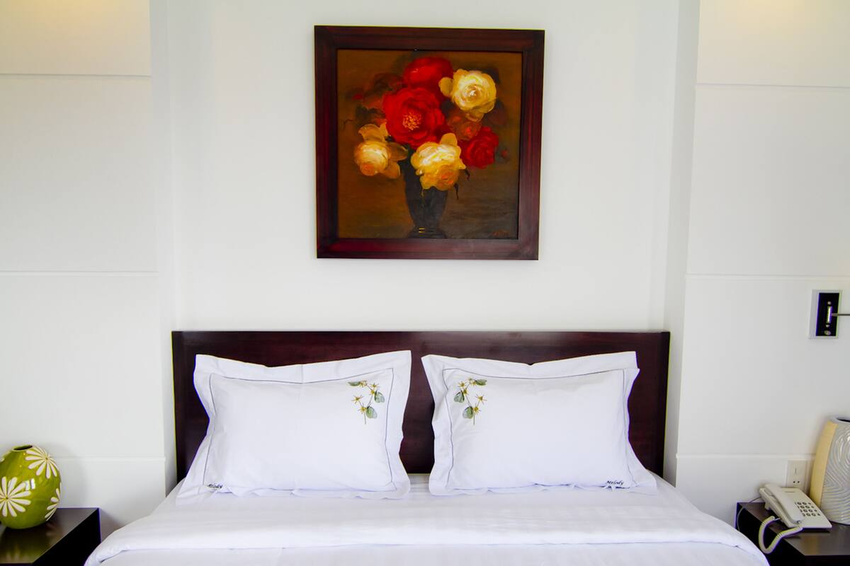 A hotel room with a bed, two pillows, a nightstand with a vase, a painting of flowers, a lamp, and a phone on the nightstand.