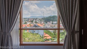 Ein Hotelzimmer mit großem Fenster, Vorhängen und Blick auf eine Stadtlandschaft mit Gebäuden und Bergen.