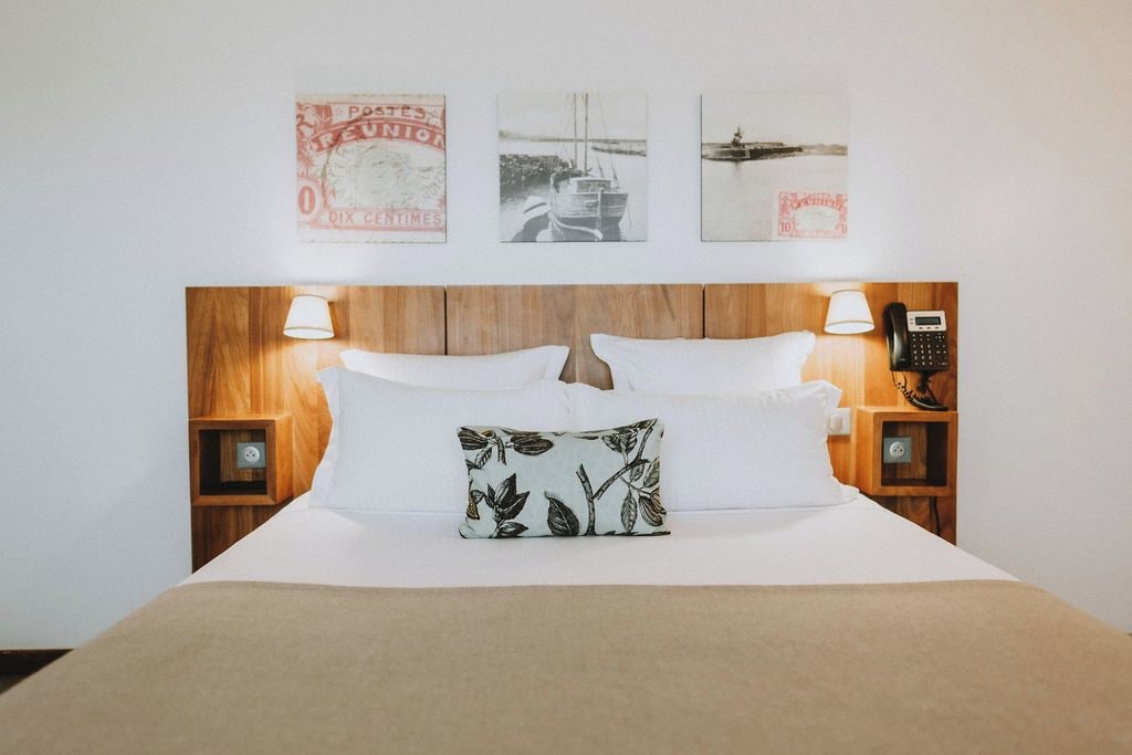 A hotel room with a wooden headboard, white bedding, a floral pillow, two bedside lamps, a telephone, and three framed pictures on the wall.