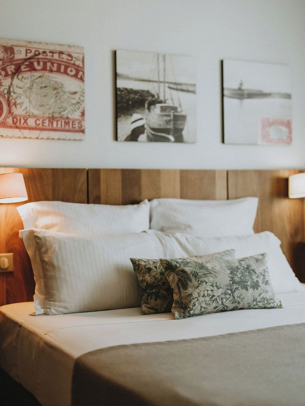 Una cama bien tendida con ropa blanca y un cojín floral, un cabecero de madera y obras de arte en la pared, incluyendo un sello postal antiguo.