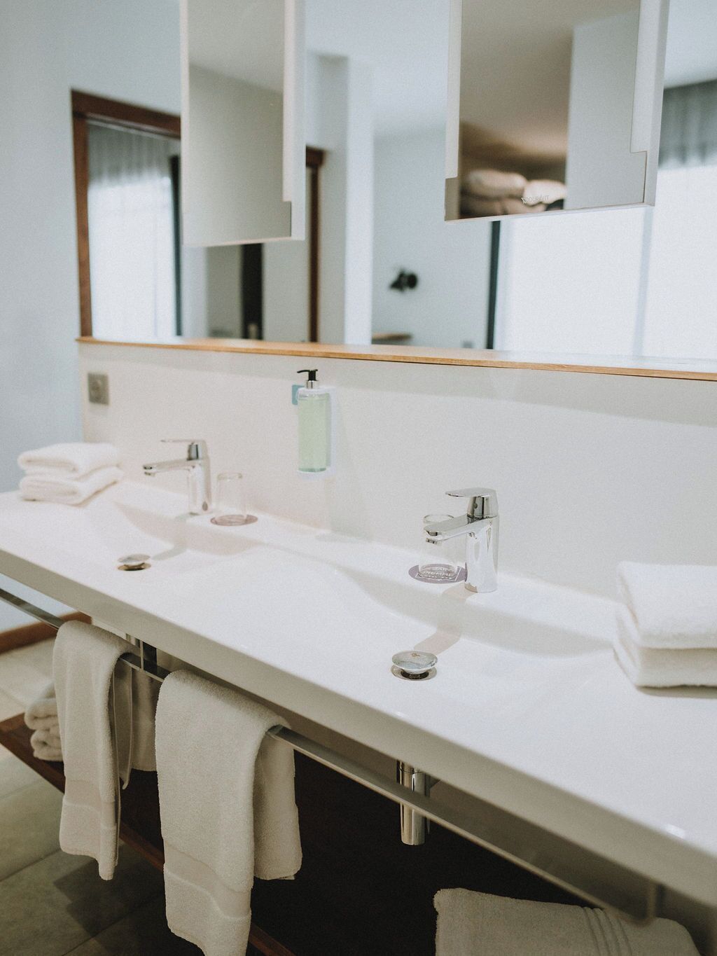 Een moderne badkamer met een lange witte wastafel, twee wasbakken, een spiegel en een handdoekhouder.