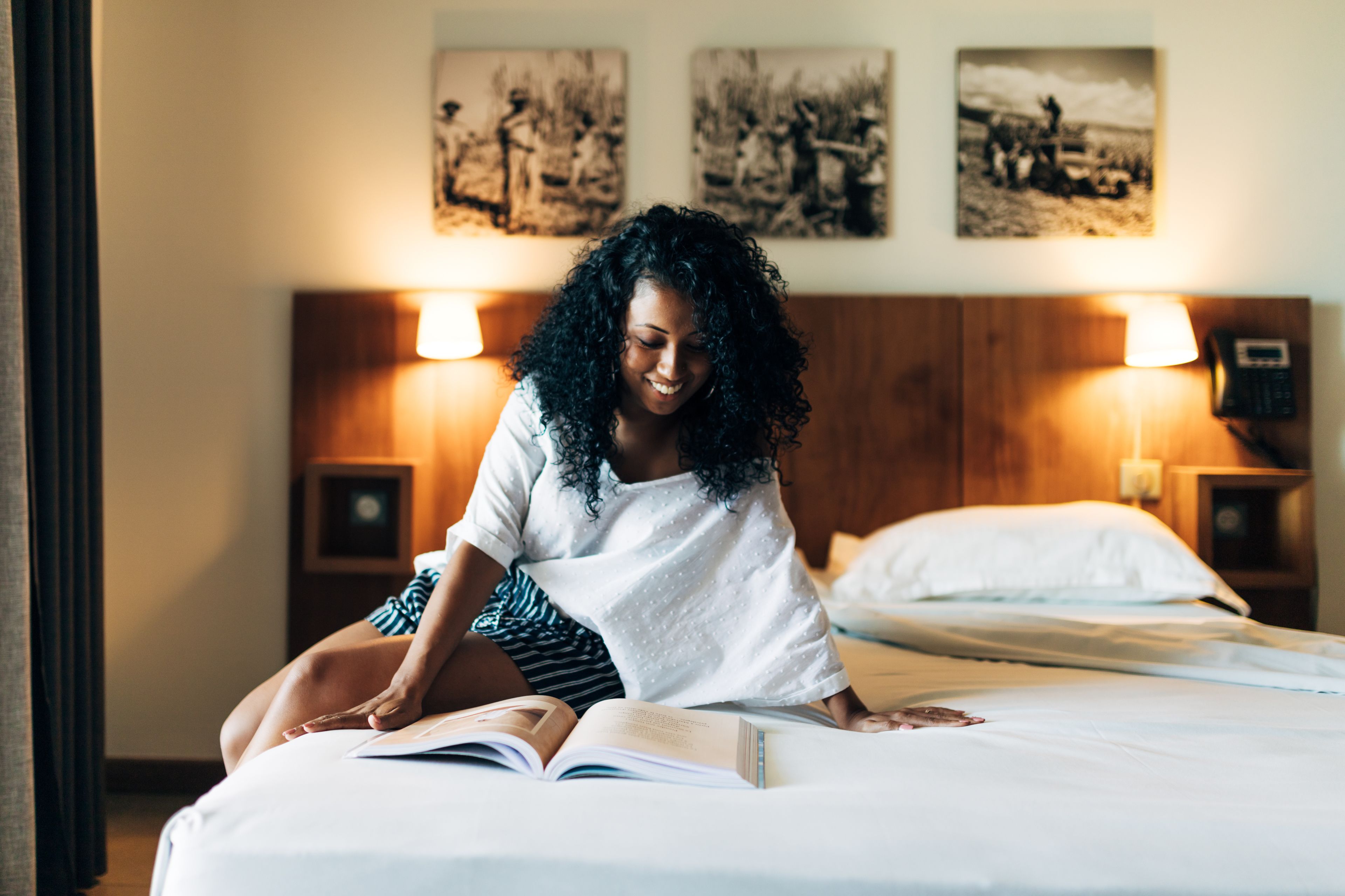 Una persona sentada en una cama con un libro abierto, en una habitación con cabecera de madera, dos lámparas de noche y cuadros en la pared.