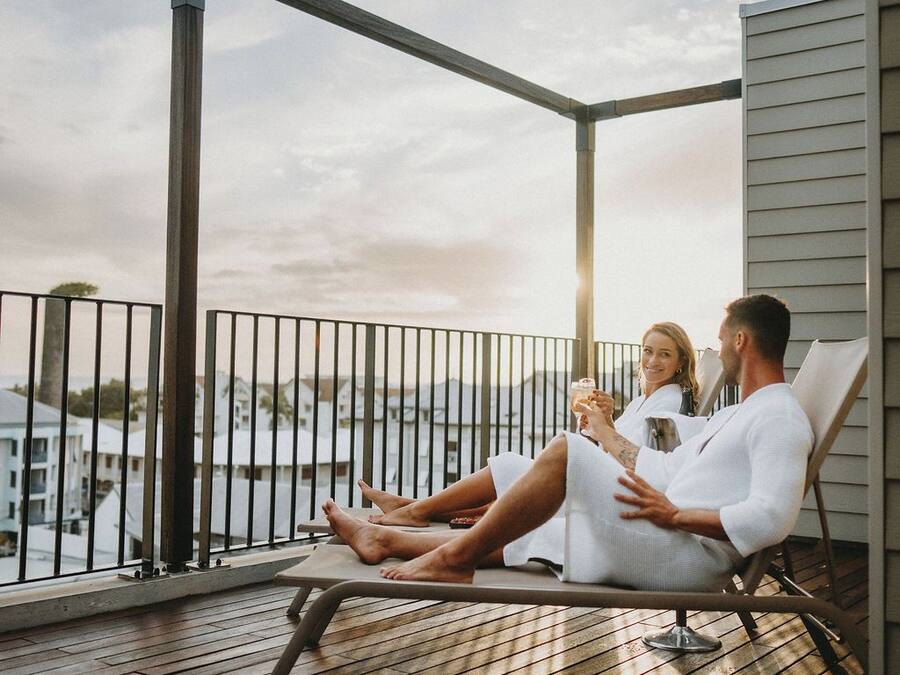 Un couple se détend sur une terrasse en bois dotée d’une rambarde en métal, avec vue sur une étendue d’eau et des bâtiments.