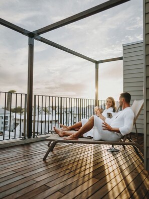 Una pareja se relaja en una terraza de madera con barandilla de metal, con vista a un cuerpo de agua y edificios.