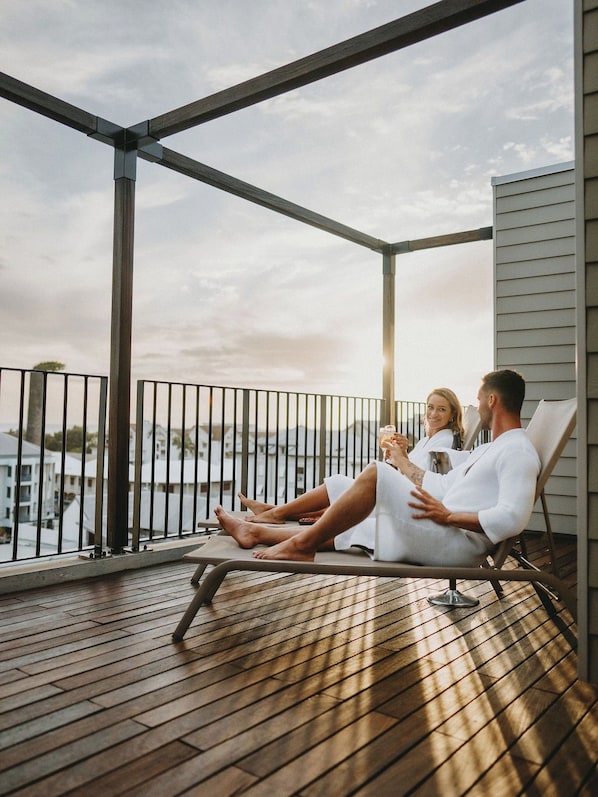 Una pareja se relaja en una terraza de madera con barandilla de metal, con vista a un cuerpo de agua y edificios.