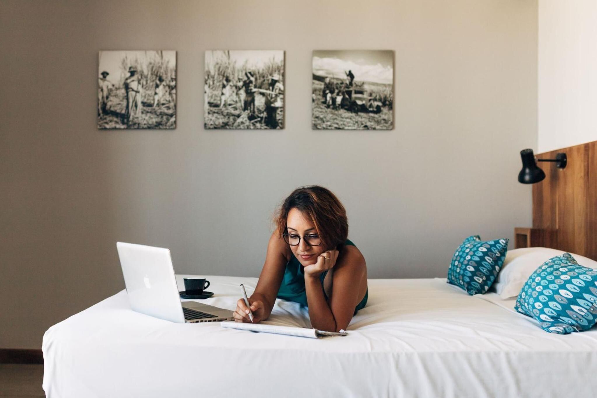 Eine Person sitzt auf einem Bett, benutzt einen Laptop und schreibt in ein Notizbuch. Dahinter hängen drei schwarz-weiße Fotografien an der Wand.