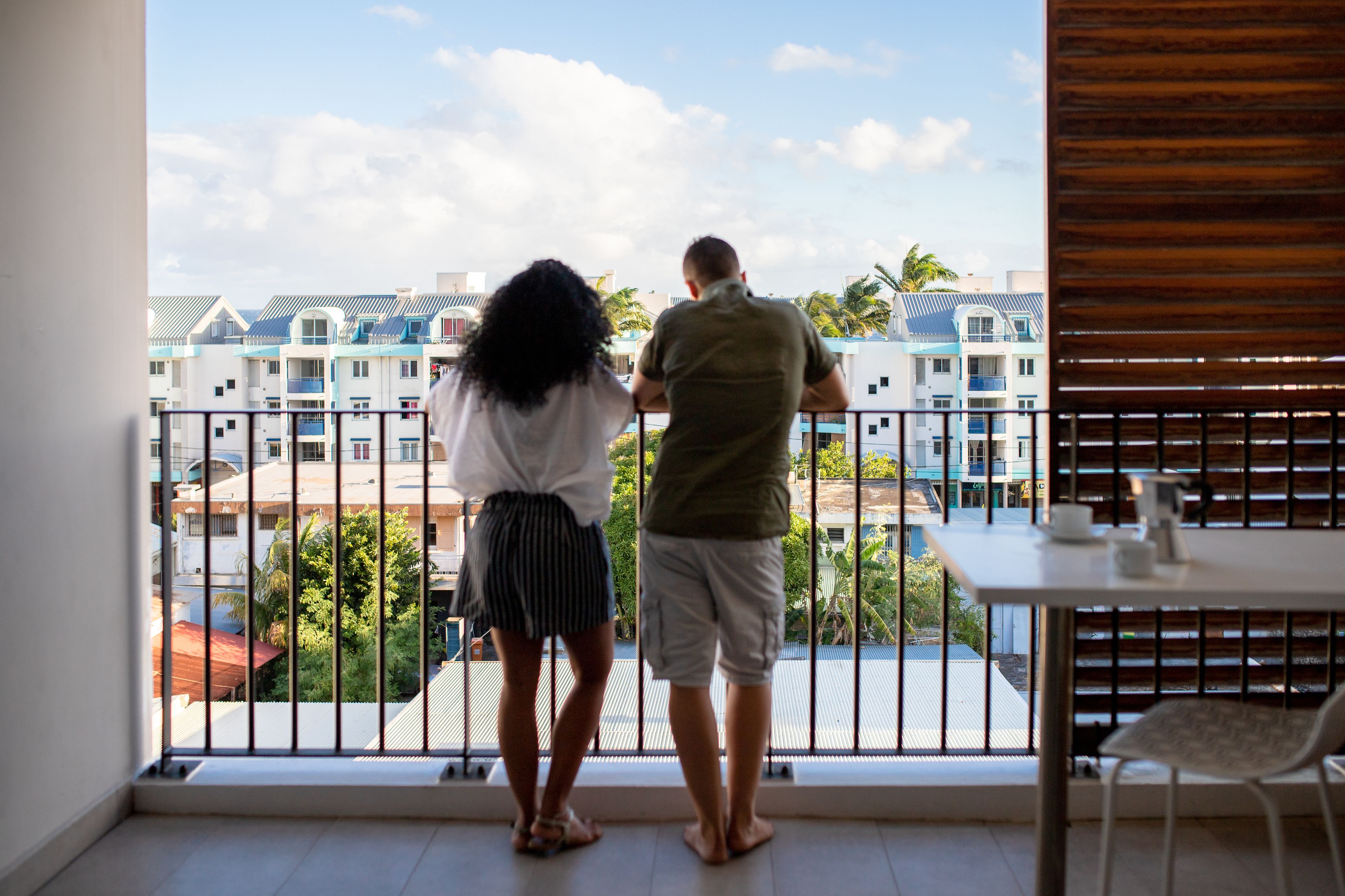 A couple standing on a balcony overlooking a residential area with multiple buildings and palm trees.