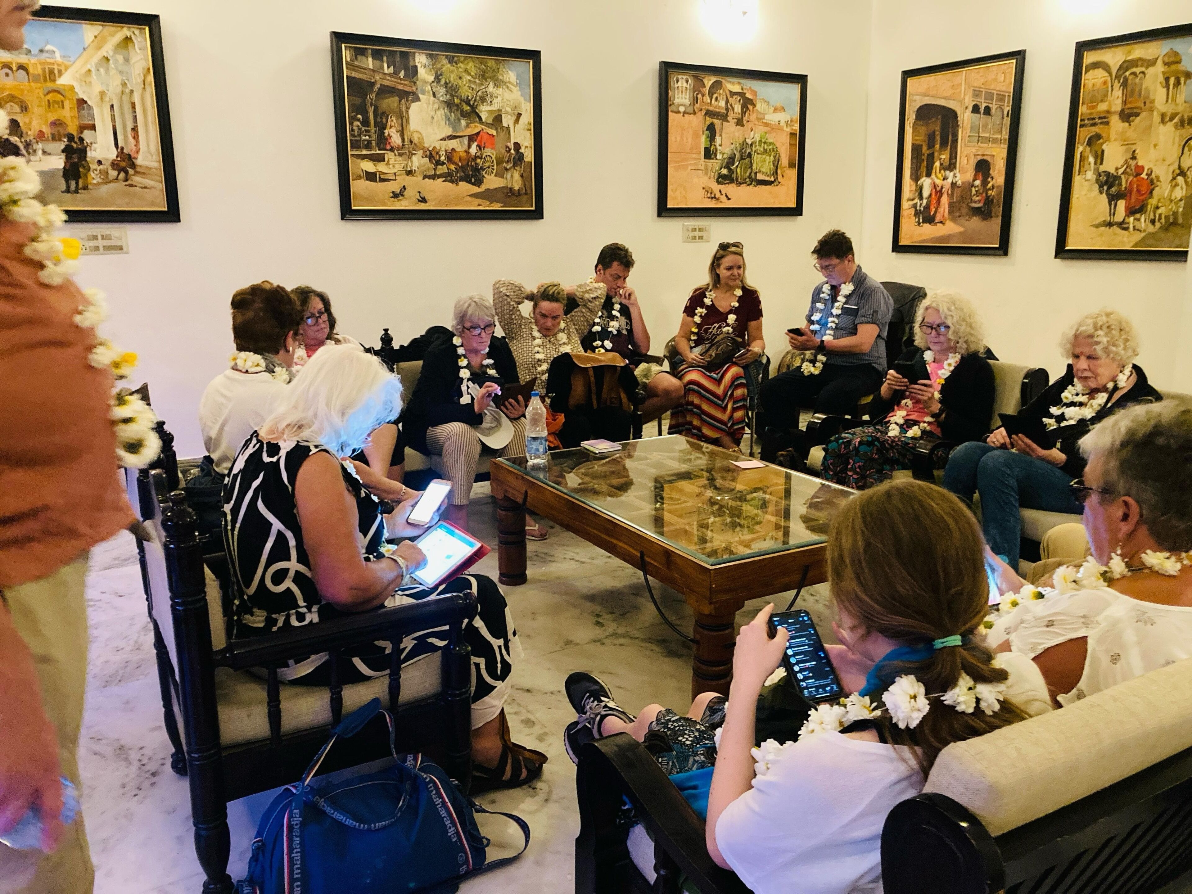 A group of people seated in a room with a glass coffee table, surrounded by framed paintings on the walls.