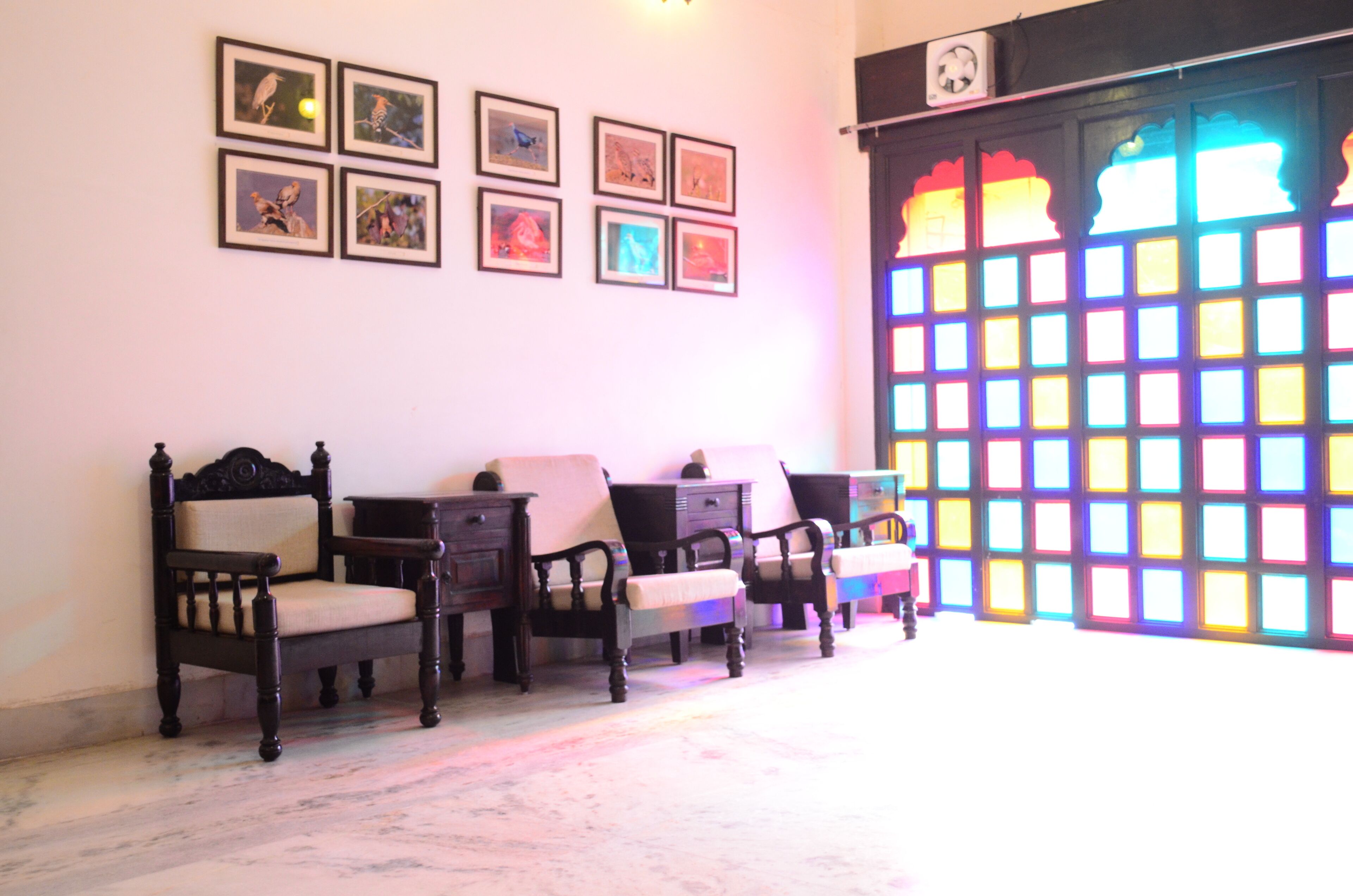 A room with a traditional wooden chair and a small table, a wall with framed pictures, and a colorful window with a grid pattern.